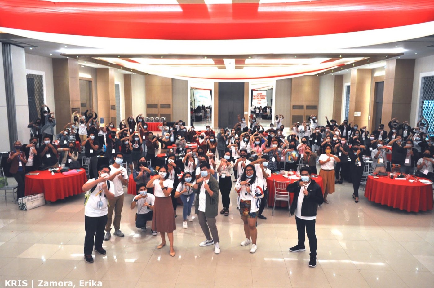 Hundreds of youth network members make a hand gesture together in a ballroom.