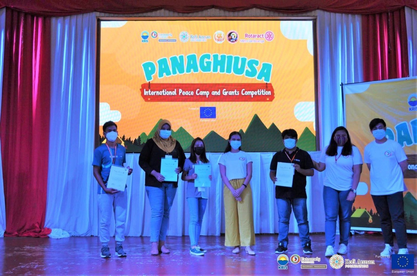 Grant winners hold their certificates on stage at the Panaghiusa Peace Camp.